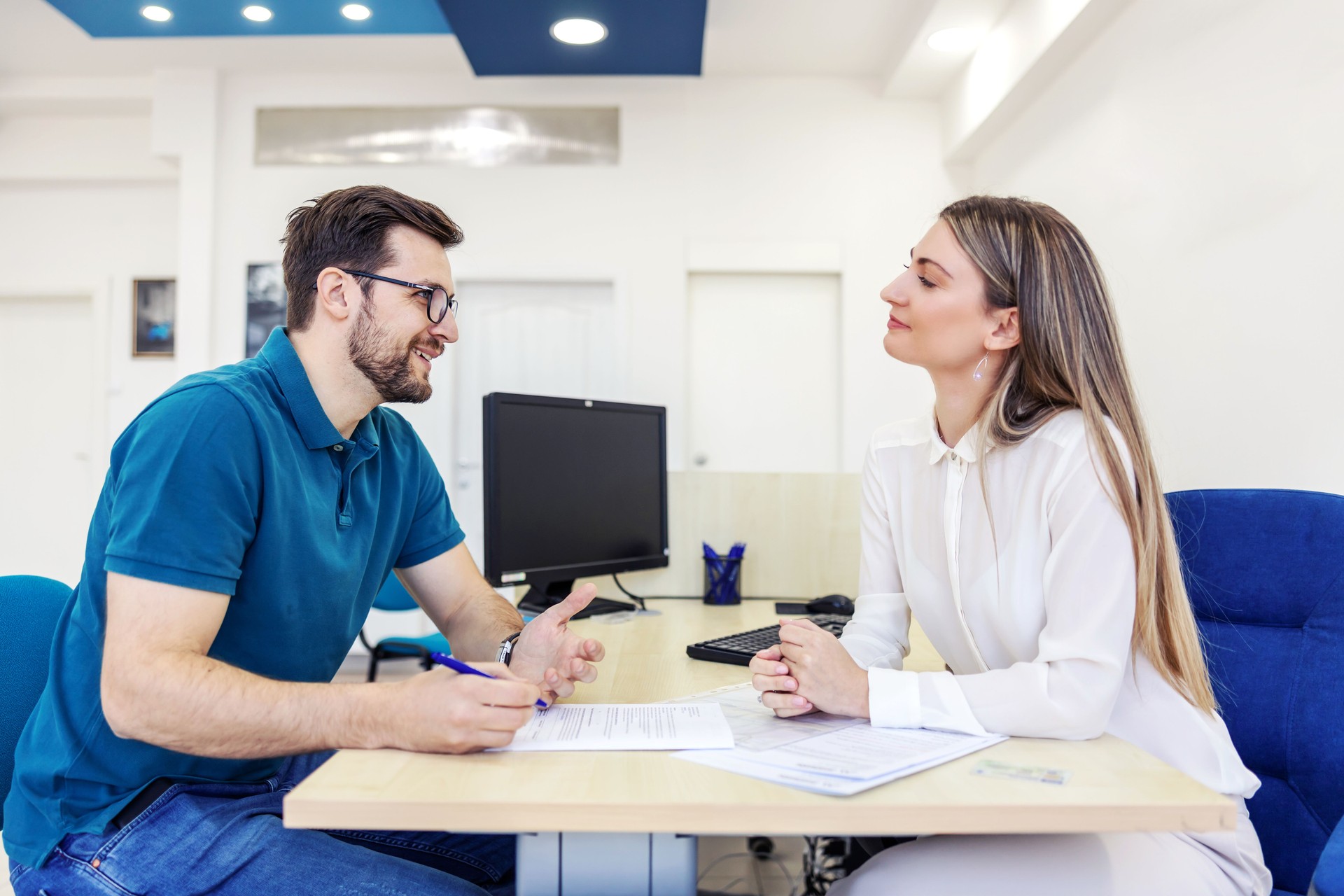 Paperwork for technical inspection. Man in casual clothes with glasses is sitting at the table of an adult woman in a car repair shop or car service. He talks to the female worker and signs the papers